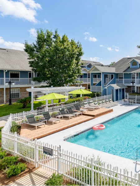 Community pool with grey lounge chairs, wooden deck, grass area, sidewalk and pergola