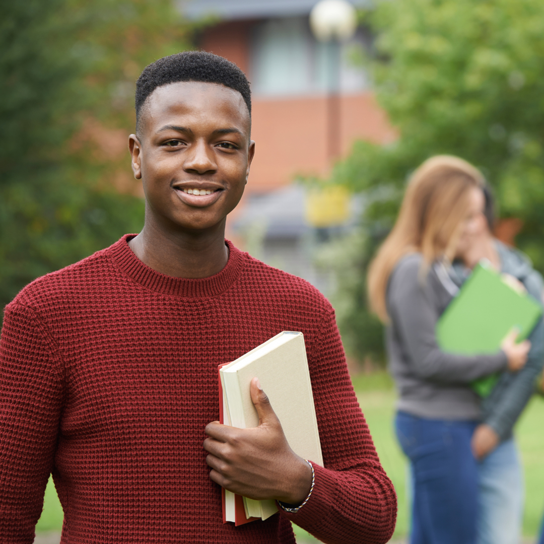 A young African American college student looks at the camera smiling. Other students are in the background talking, but blurred.