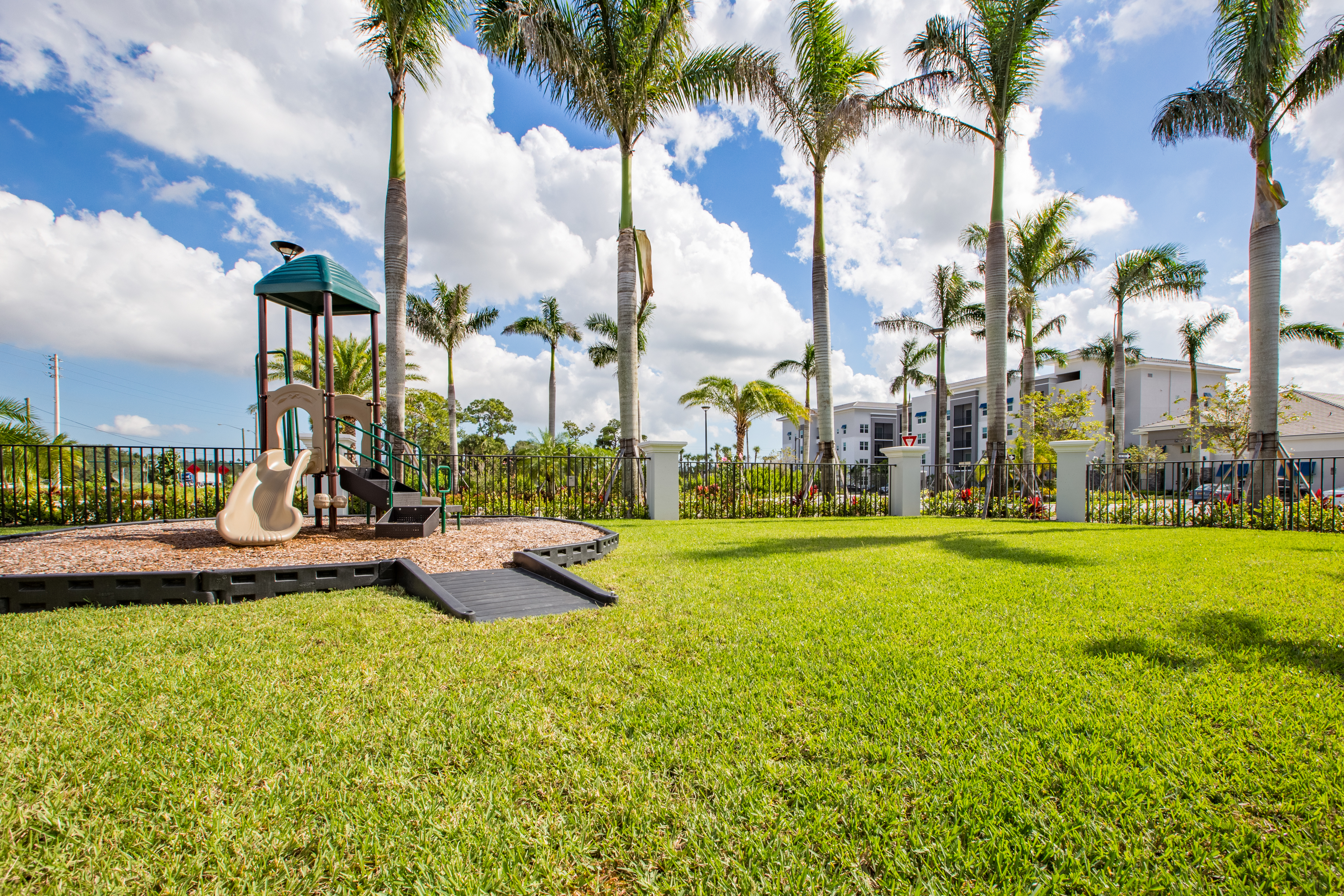 View of Playground, Showing Slide, Climbing Structure, Fenced-In, and Palm Trees in Background at Cottonwood West Palm Apartments