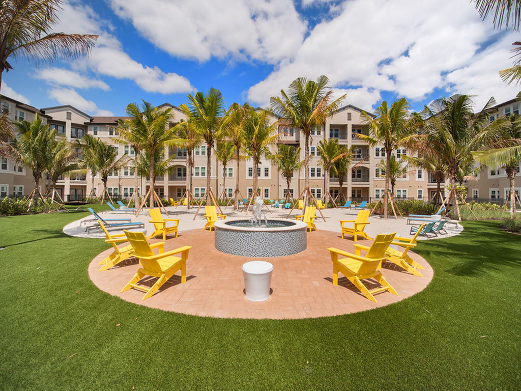 View of Seating Near Water Feature, Showing Water Feature Circled with Seating Near Grass in Courtyard at Murano at Three Oaks Apartments